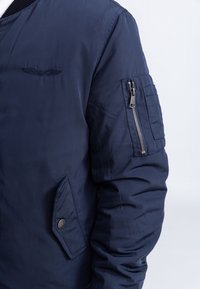 Personne portant un blouson aviateur bleu marine avec une poche zippée sur la manche, une poche avant en biais et un petit logo brodé sur la poitrine.