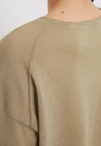 Close-up van de bovenrug en schouder van een persoon die een beige gebreide trui draagt met zichtbare stikseldetails.