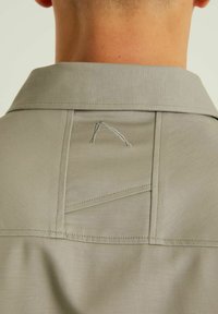 Šviesiai pilka marškinė su apykakle, pagaminti iš tekstūruotos medžiagos, turi siuvinėtą kišenę ir minimalistinį siuvinėjimo elementą nugaros pusėje.