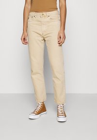 Jeans de mezclilla beige con corte recto, diseño de cinco bolsillos y dobladillo deshilachado. Combinados con zapatillas altas marrones.