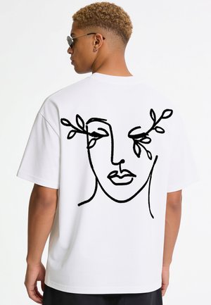 Jonge man met kort krullend haar, een zonnebril en een wit T-shirt met een zwarte lijnttekening van een gezicht met takken als wenkbrauwen.