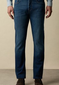 Tmavě modré džíny s rovnými nohavicemi, se standardními kapsami a jemným vyblednutím, spojené s hnědými botami na neutrálním pozadí.