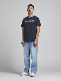 Tamsiai mėlynas marškinėliai su baltu "JACK & JONES" logotipu, derinami su šviesiai mėlynomis laisvo kirpimo džinsomis ir baltais sportiniais bateliais. Pilno kūno vaizdas.