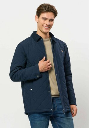 Jeune homme souriant, portant une veste matelassée bleu marine sur un pull beige et un jean bleu, debout devant un fond clair uni.