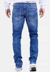 Jean en denim bleu à coupe slim, avec plusieurs poches arrière et des détails de couture. Porté avec des baskets blanches.
