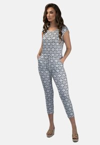 Biały kombinezon z czarnym geometrycznym wzorem; krótkie rękawy, elastyczna talia, boczne kieszenie i zwężane nogawki. Beżowe sandały na koturnie dopełniają stylizację.