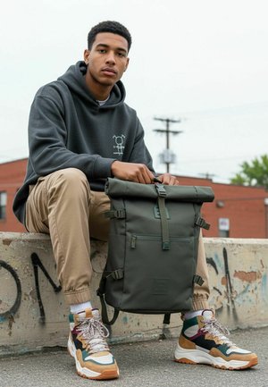 Un giovane uomo con una felpa con cappuccio scura e pantaloni color sabbia è seduto su un muretto di cemento tenendo uno zaino verde scuro con chiusura arrotolabile all'aperto.