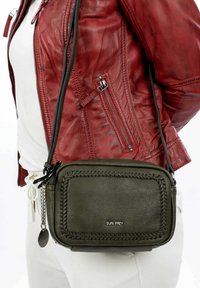 Zelená kožená crossbody taška s pletenými okraji, stříbrným kováním a odnímatelným řetízkem, nošená přes červenou koženou bundu.