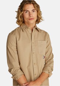 Camicia beige a maniche lunghe con bottone frontale, colletto classico, taschino singolo sul petto e logo ricamato sulla tasca. Tessuto di cotone morbido.