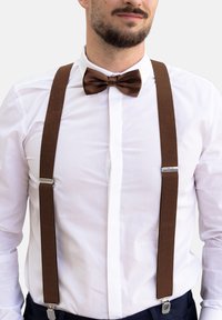Tirantes de tela marrón con clips plateados combinados con una pajarita marrón a juego, llevados sobre una camisa blanca de vestir. Diseño y textura simples.