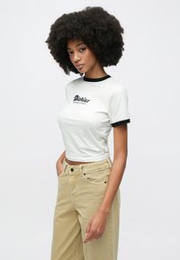 T-shirt blanc à manches courtes avec col contrastant noir, arborant le logo texte "Dickies" en noir. Le modèle porte un pantalon taille haute beige clair.