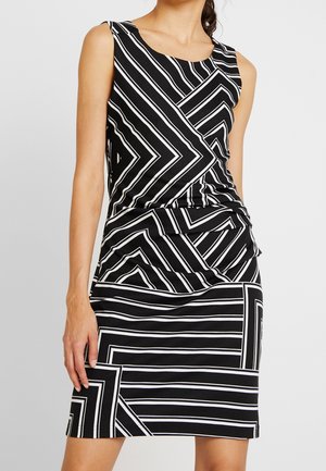 Hiatonen musta mekko, jossa valkoisia geometrisia chevron- ja raidakuoseja, istuva vyötärö, päällä seisovalla henkilöllä, jonka toinen käsi lepää sivulla.