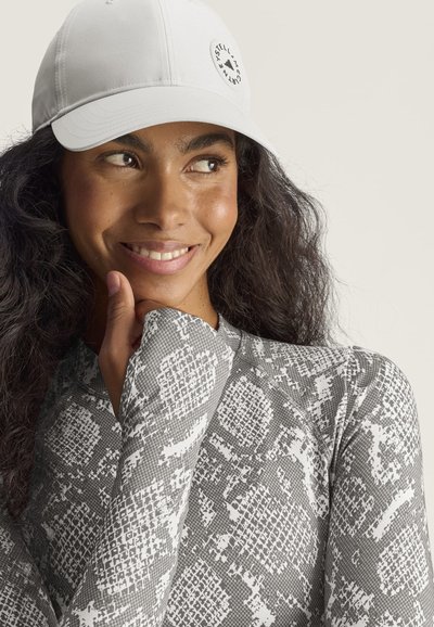 Mujer con cabello largo y oscuro, que lleva una gorra blanca y una camiseta gris de manga larga con estampado, sonriendo y mirando hacia un lado con la mano cerca del mentón.