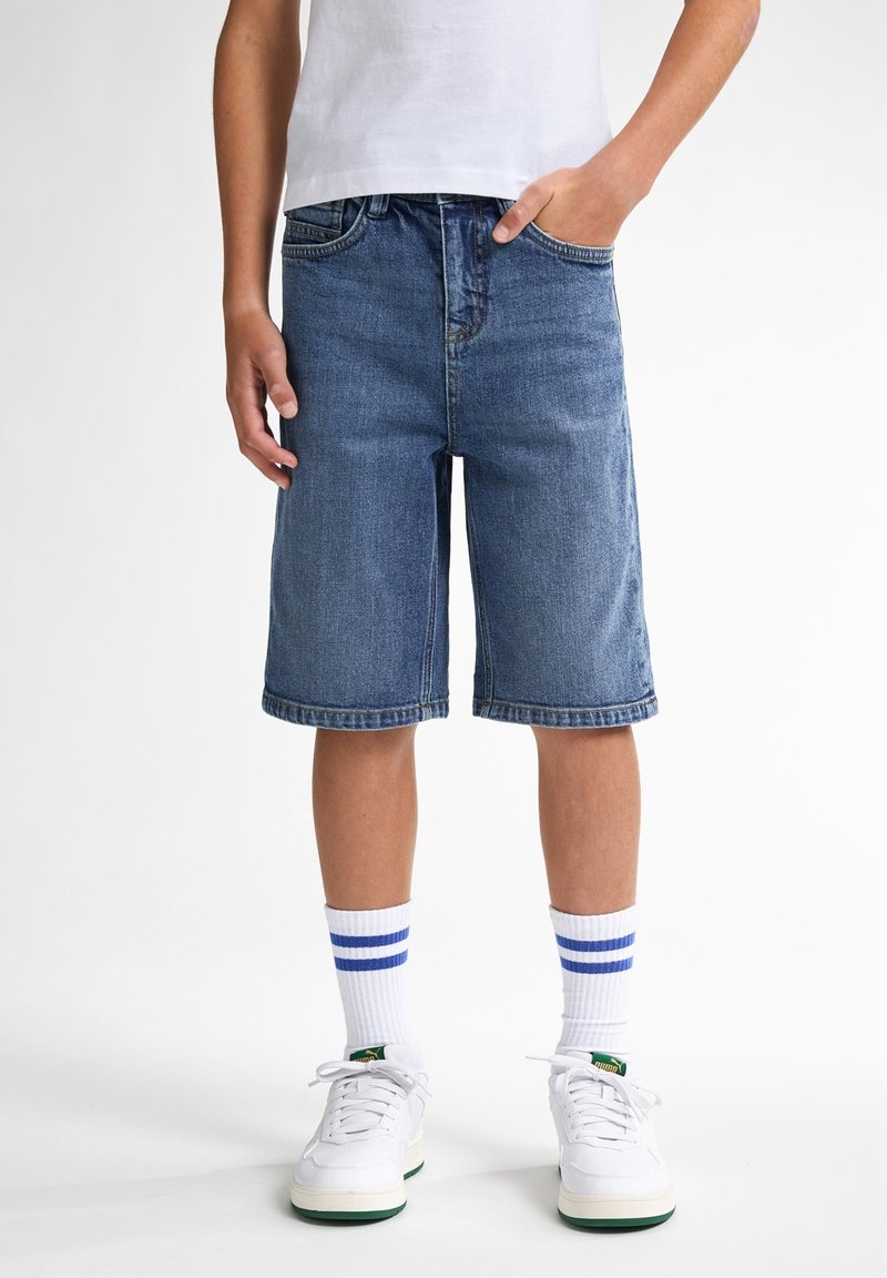 Jeansshorts i blått med rak passform, har fem fickor och upprullad nederkant, kombinerat med vita sneakers och randiga strumpor.