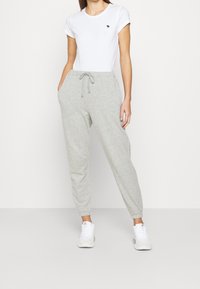 Harmaat collegehousut, joissa on joustava resori nilkoissa ja kiristettävä vyötärö, yhdistettynä tiukkaan valkoiseen T-paitaan, jossa on pieni logo rinnassa.