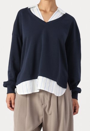 Osoba ubrana w warstwowe ubrania: granatowy sweter z dekoltem w serek na białej koszuli w niebieskie paski oraz beżowe plisowane spodnie.