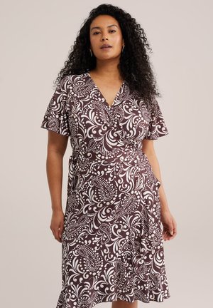 Nő hosszú, göndör hajjal, barna és fehér virágmintás, rövid fodros ujjú csavart ruhát viselve, sima háttér előtt áll.