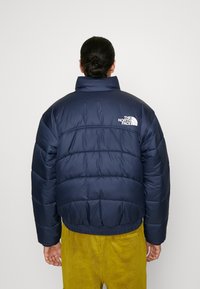 Marinblå puffjacka med horisontell quiltning, hög krage och elastisk midja. Har en vit logotyp på övre delen av ryggen.