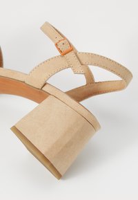 Sandalia de tacón cuadrado beige con correas cruzadas estrechas, que presenta una pequeña hebilla naranja y una textura parecida a la gamuza. La base es de madera.