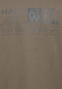Brun t-shirt med blå tryckt text som inkluderar "HALO", "OFF DUTY GEAR" och andra detaljer; mjuk textur med minimal lyster.