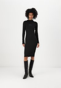 Vestido bodycon preto com mangas longas e gola alta. Apresenta um logótipo discreto e um comprimento até à meia panturrilha, combinado com botas pretas brilhantes até ao joelho.