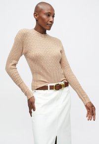 Pulover beige din tricot cu model in dungi, cu maneci lungi și decolteu rotund, purtat împreună cu o fustă albă și o curea maro cu o cataramă mare aurie.