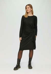 Schwarzes knielanges Kleid mit langen Ärmeln, das ein gedrehtes Design und strukturierten Stoff aufweist. Kombiniert mit schwarzen Knöchelstiefeln.
