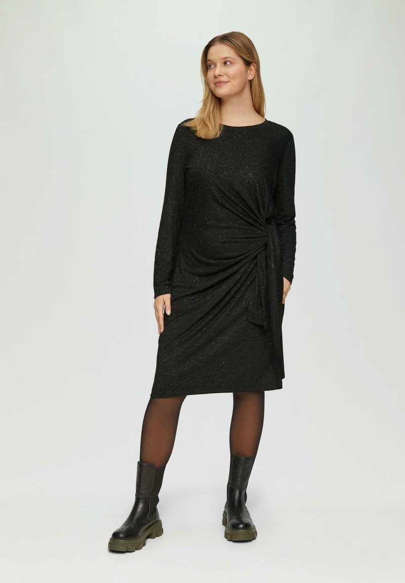 Schwarzes knielanges Kleid mit langen Ärmeln, das ein gedrehtes Design und strukturierten Stoff aufweist. Kombiniert mit schwarzen Knöchelstiefeln.