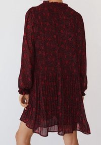 Punainen ja musta kukkakuosinen mekko, jossa on pleatattu muotoilu, pitkät hihat ja röyhelökaulus, tarjoten virtaavaa siluettia ja pehmeää tekstuuria.