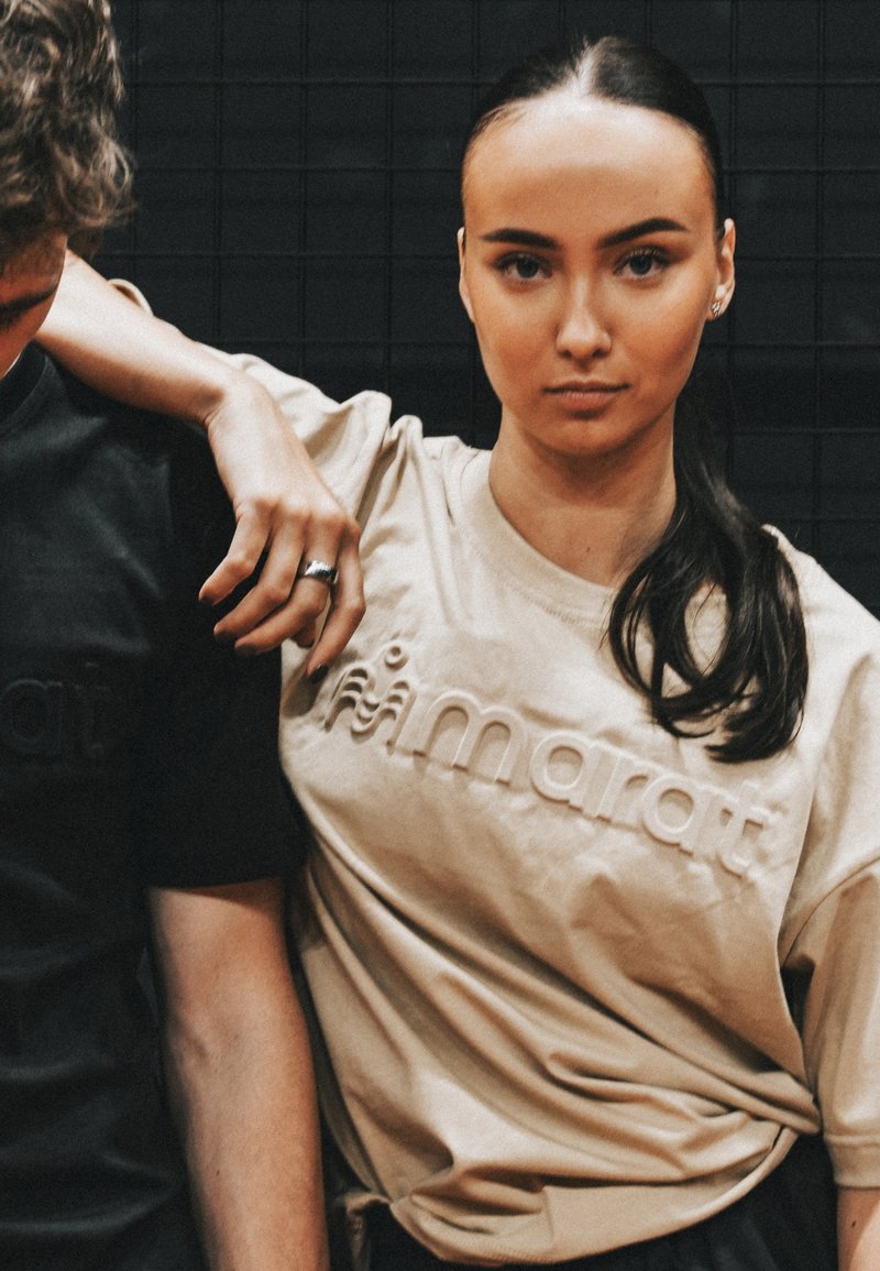 Noor naine tumedate juustega, beezivärvilises T-särgis, toetub musta T-särgiga inimesele tumeda plaatidega seina taustal.