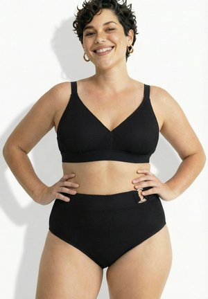 Glunderende vrouw met kort krullend haar, gekleed in een zwarte, gestructureerde tweedelige zwemoutfit met hooggetailleerde broekjes en gouden oorringen, handen op de heupen.