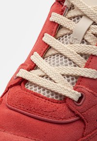 Red suede sneaker with thick cream braided laces over white mesh fabric in detailed close-up.