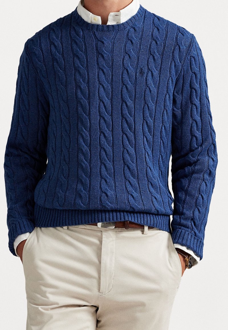 Homme portant un pull en maille torsadée bleu sur une chemise blanche à col, pantalon beige et ceinture marron, mains partiellement dans les poches.