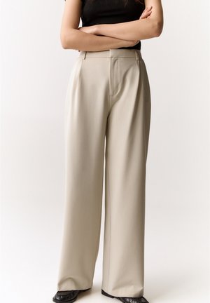 Femme bras croisés portant un haut noir sans manches, un pantalon beige à jambes larges et des chaussures noires, debout devant un fond uni.