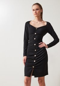 Robe noire à manches longues avec un décolleté carré, en tissu froncé, fermeture à boutons sur le devant et fente sur le côté, fabriquée dans un matériau lisse.