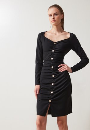 Robe noire à manches longues avec un décolleté carré, en tissu froncé, fermeture à boutons sur le devant et fente sur le côté, fabriquée dans un matériau lisse.