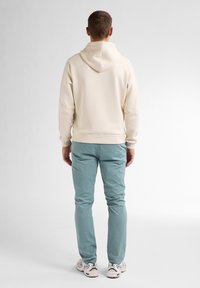 Cremefarbener Hoodie mit einer Naht auf der Rückseite, kombiniert mit hellblauen Hosen. Das Outfit verfügt über elastische Bündchen und eine lockere Passform. Weiße Sneakers sind sichtbar.