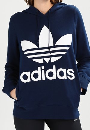 Personne portant un sweat à capuche bleu marine avec grand logo blanc Adidas et texte, mains dans les poches, associé à un pantalon noir.