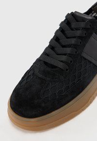 Sneaker en daim noir avec des détails en maille, bout rond, semelle épaisse en caoutchouc gomme et lacets plats noirs. Présente des accents texturés sur les côtés.
