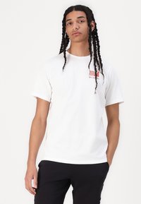White cotton t-shirt with short sleeves, featuring a red graphic logo on the chest. Simple, relaxed fit, worn with black pants.