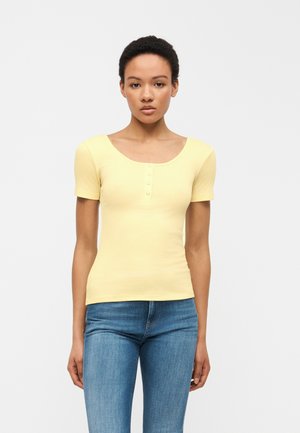 Γυναίκα με κοντά σγουρά μαλλιά, ντυμένη με ανοιχτόκίτρινη μπλούζα με κοντά μανίκια και μπλε τζιν, που στέκεται μπροστά σε ένα απλό φωτεινό φόντο.