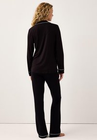 Donna con capelli biondi ricci che indossa un pigiama nero a maniche lunghe con piping bianco, in piedi a piedi nudi su uno sfondo semplice.
