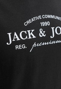 Μαύρο ύφασμα με λευκό τυπωμένο κείμενο: «Creative Community 1990 JACK & JONES REG. premium» σε ανάμεικτες γραμματοσειρές και στυλ.