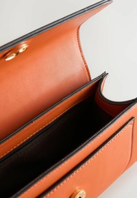 Orange läderväska med en slät yta, som har en svart foder, en dragkedjeficka och en guldfärgad snapstängning. Insidan har två fack.