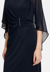 Granatowa sukienka z gładkiego materiału, z marszczeniami w pasie i przezroczystymi, falującymi rękawami. Posiada dekoracyjny akcent w postaci cyrkonii.
