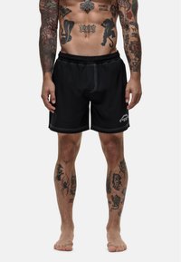 Zwarte zwembroek met witte stiksels, elastische tailleband en zonder zichtbare graphics, gedragen door een persoon met zichtbare tatoeages.