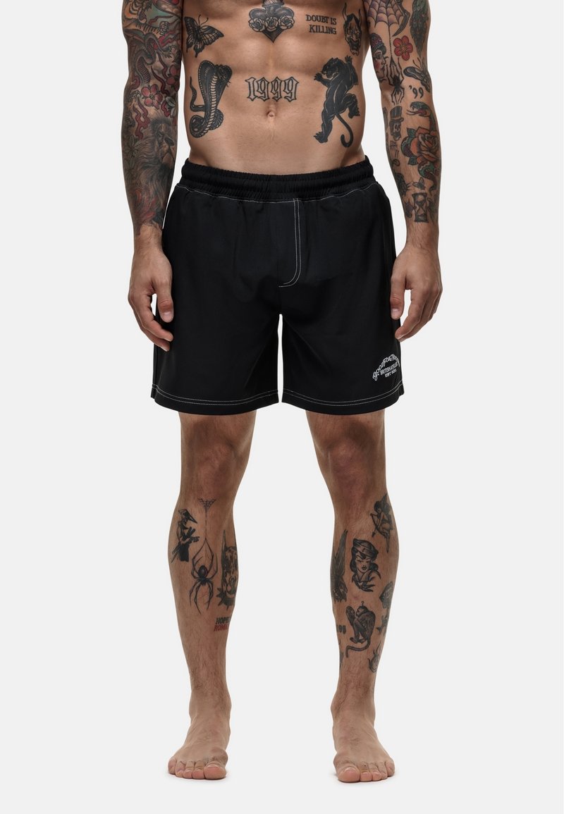 Zwarte zwembroek met witte stiksels, elastische tailleband en zonder zichtbare graphics, gedragen door een persoon met zichtbare tatoeages.