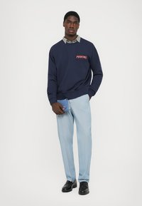 Felpa navy con logo "MARNI", abbinata a pantaloni leggeri a righe verticali blu chiaro. Il modello tiene in mano una piccola borsa blu e indossa scarpe nere.