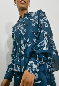 Modrá květinová košile s předním zapínáním na knoflíky, dlouhými rukávy se staženými manžetami a hladkou texturou. Vzor obsahuje bílé a tmavě modré akcenty.