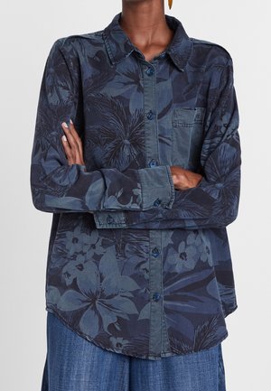 Person wearing a long-sleeve navy blue floral button-up shirt with pocket, arms crossed, paired with loose blue pants.